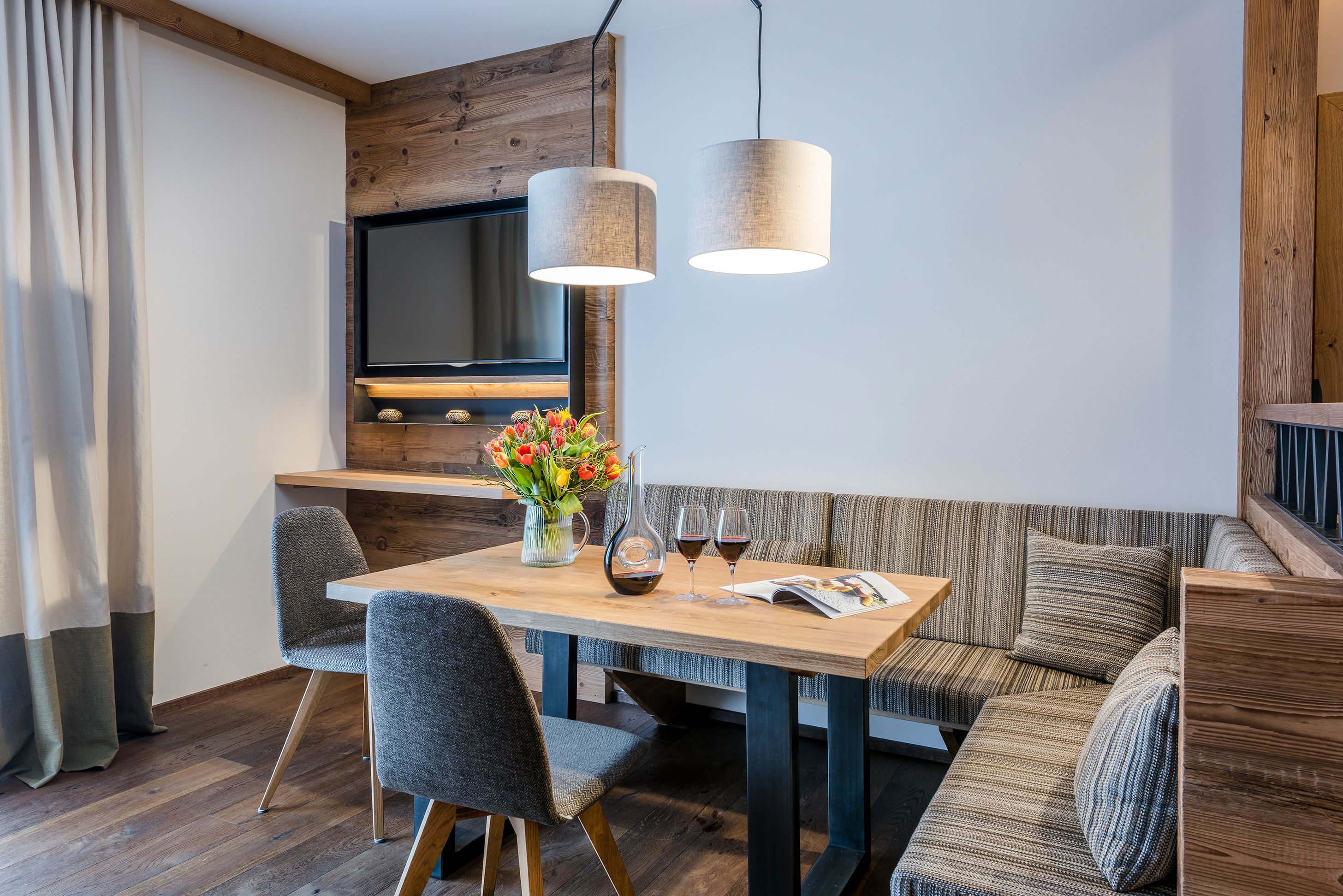 A modern living room with a wooden dining table, two grey chairs, a sofa, and a TV mounted on a wooden wall.