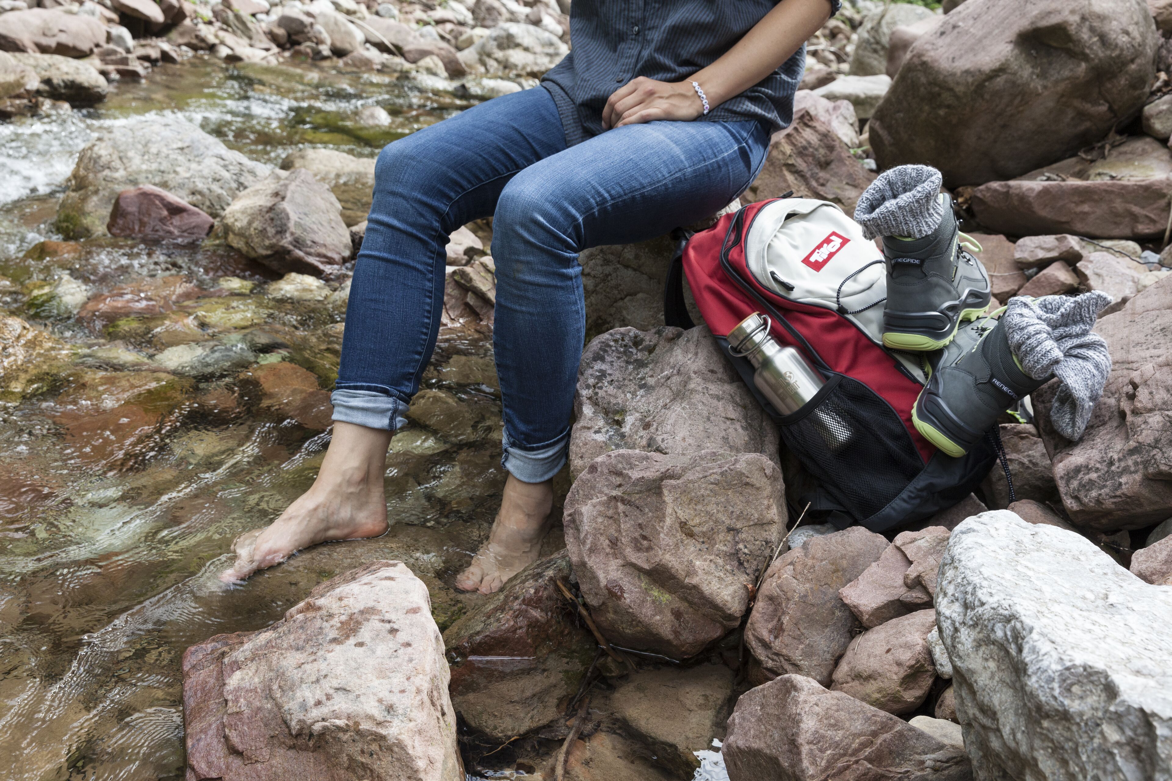 Eine Person sitzt auf Felsen an einem Bach, trägt Jeans und eine Armbanduhr.