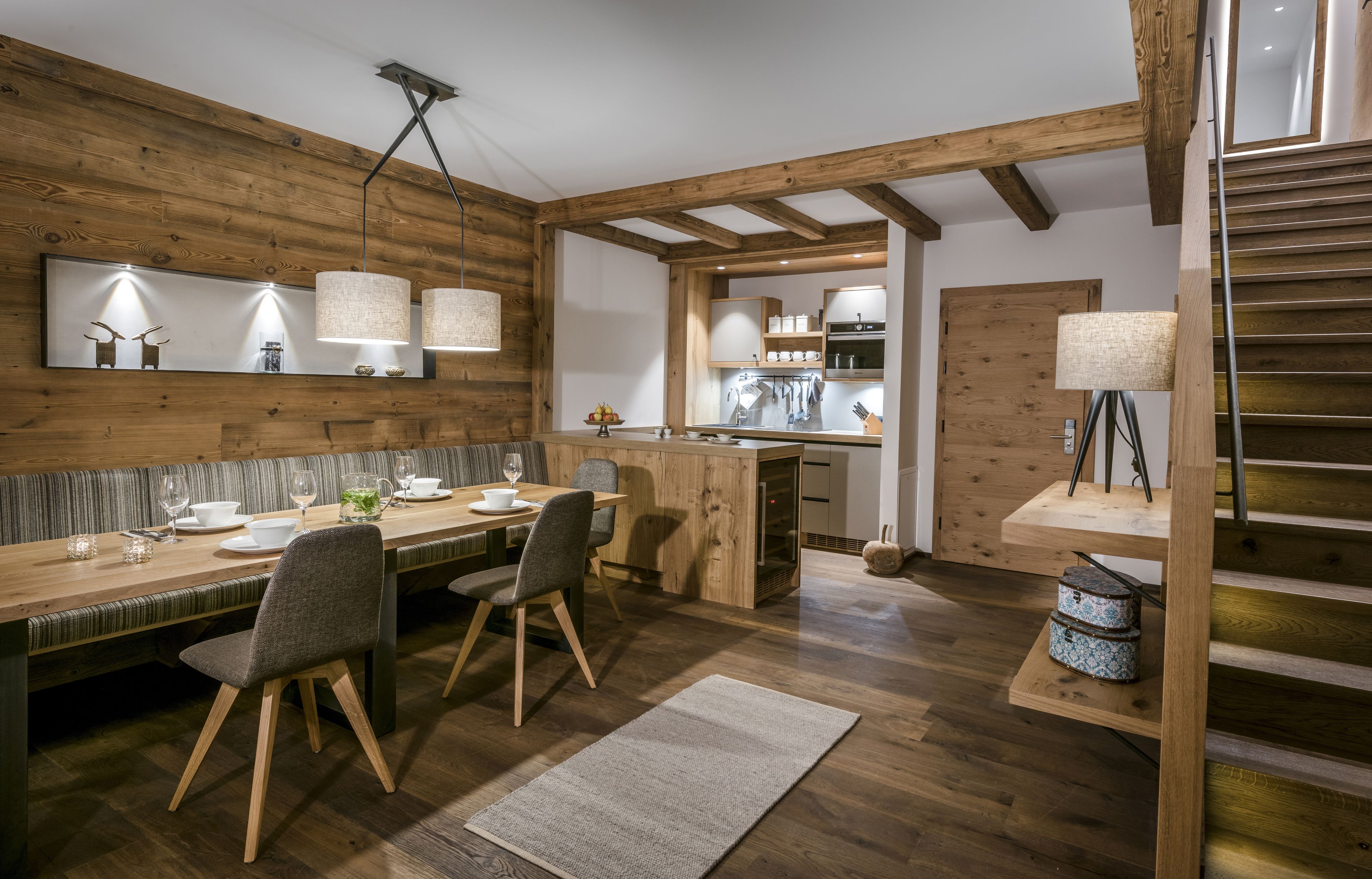 A modern kitchen with wooden beams, a dining area with a long table and chairs, and a staircase with a lamp.