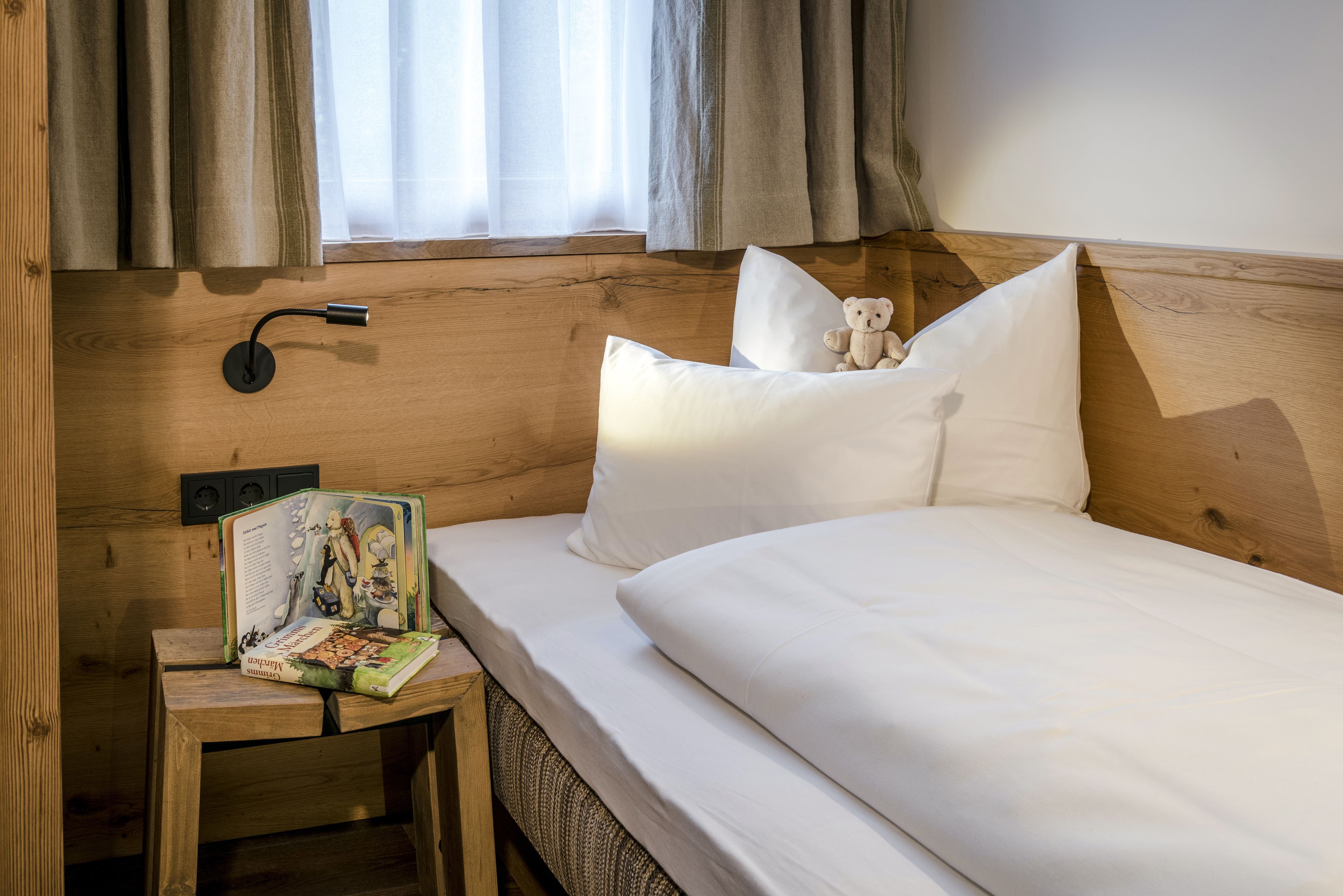 A neatly made bed with white linens, a small bedside table with a book and a teddy bear, and a wall-mounted lamp.
