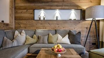 Un salon moderne avec un plafond en bois, un canapé gris agrémenté de plusieurs coussins, une table basse en bois sur laquelle repose un panier de fruits, et un lampadaire.