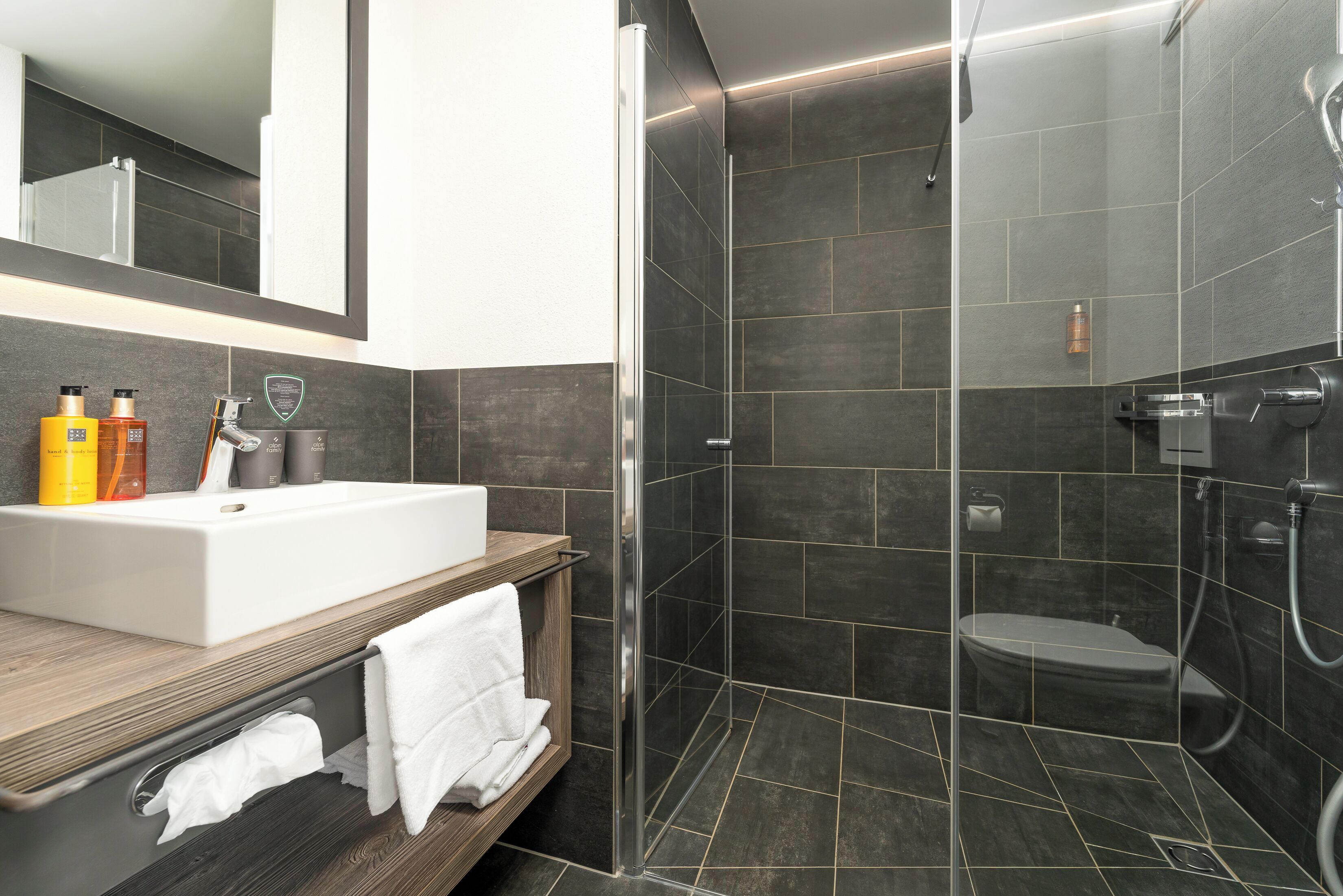 Ein modernes Badezimmer mit einer gläsernen Duschkabine, einem weißen Waschbecken und dunkel gefliesten Wänden.
