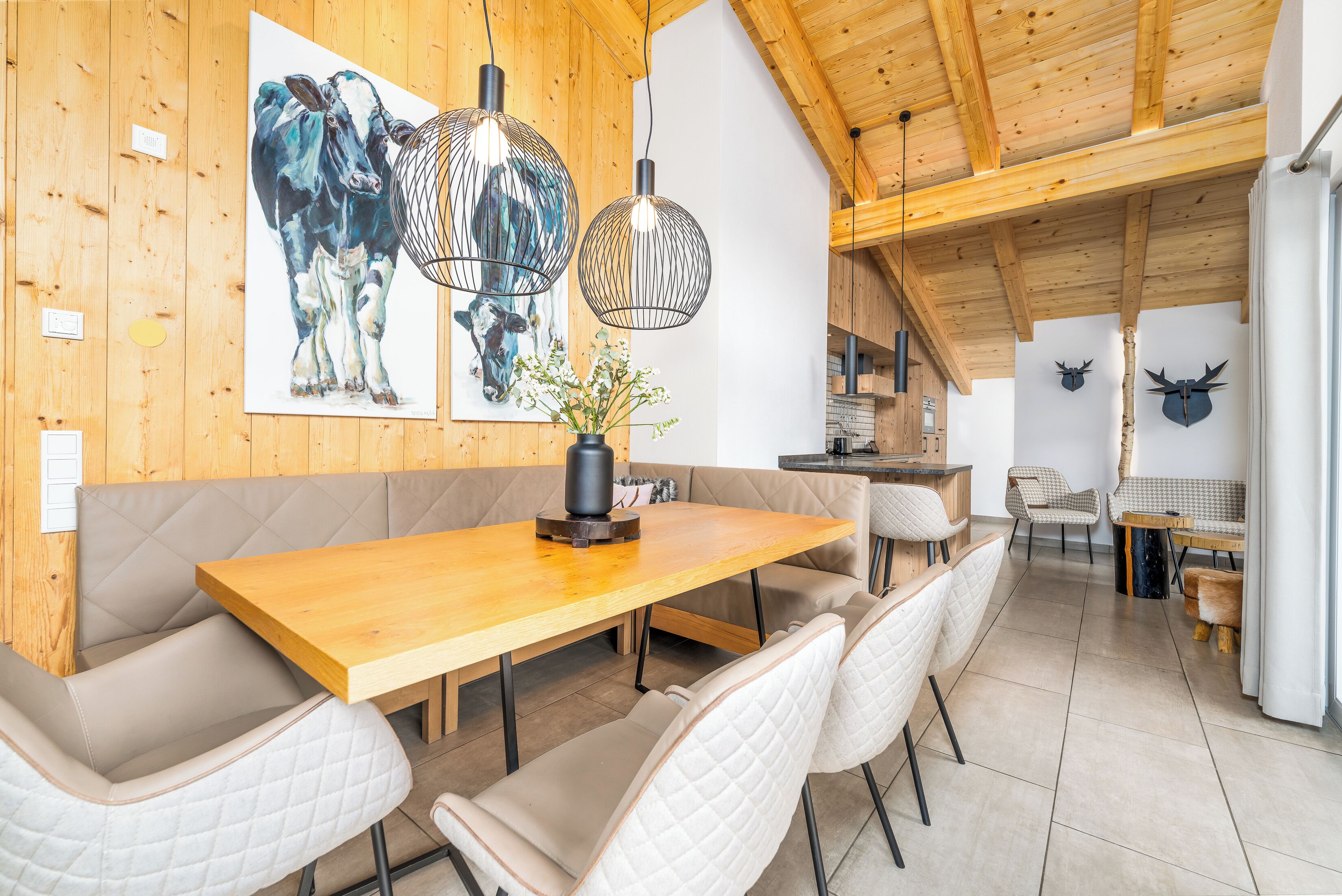 A dining area with a wooden table and cushioned benches, a painting of a cow, and two hanging light fixtures.