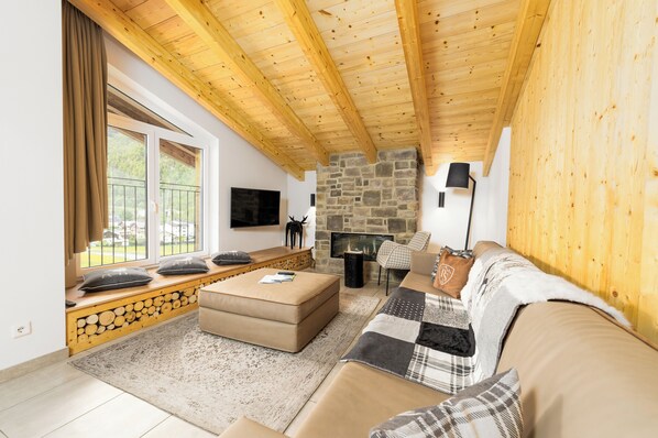 A cozy living room with a stone fireplace, wooden ceiling beams, a large window with curtains, and a comfortable seating area. - AvenidA Panorama Suites Kaprun (Kaprun)