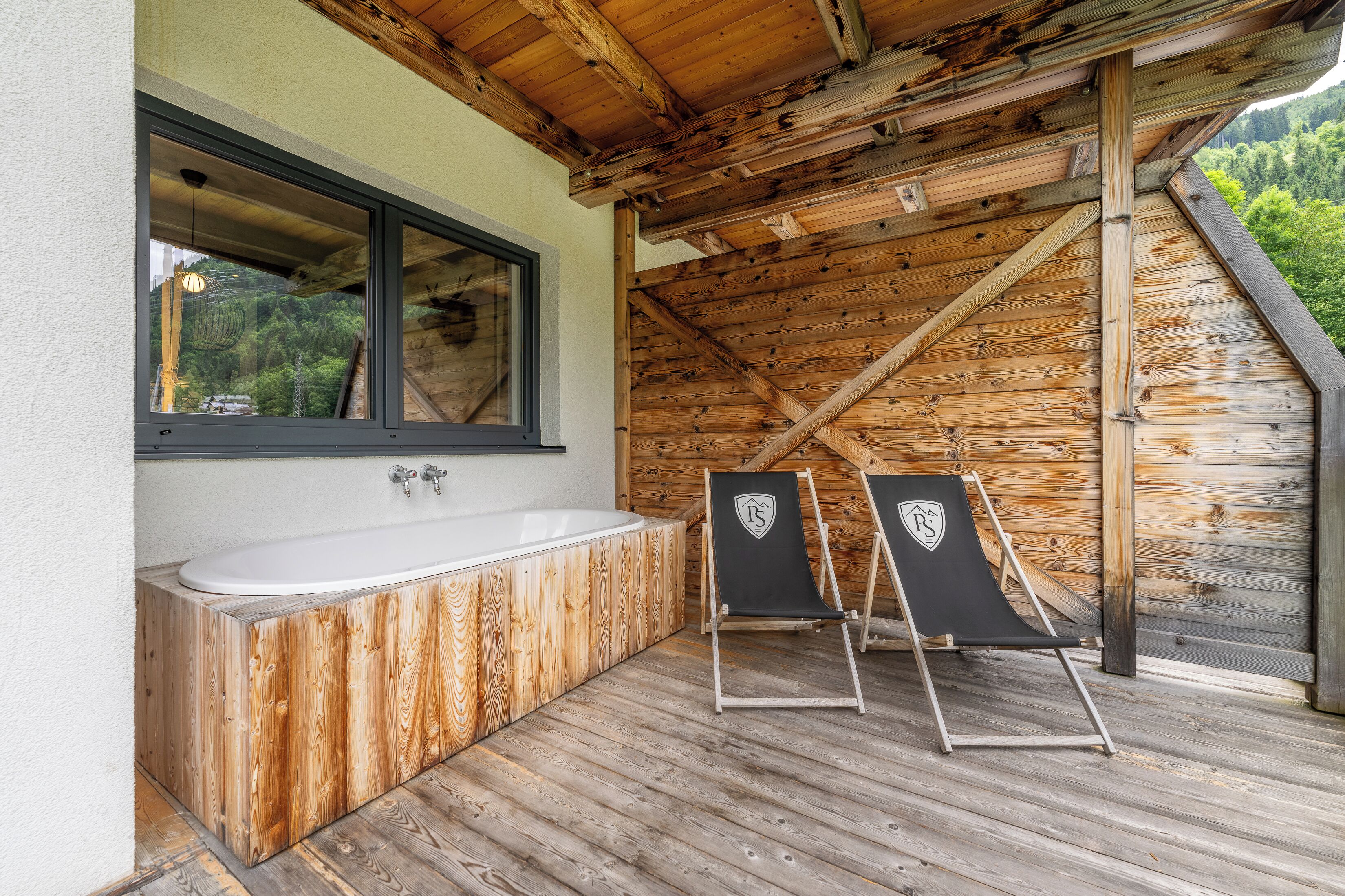 Una cabaña de madera con una bañera de madera, dos sillas de playa y una ventana con vistas a un bosque.