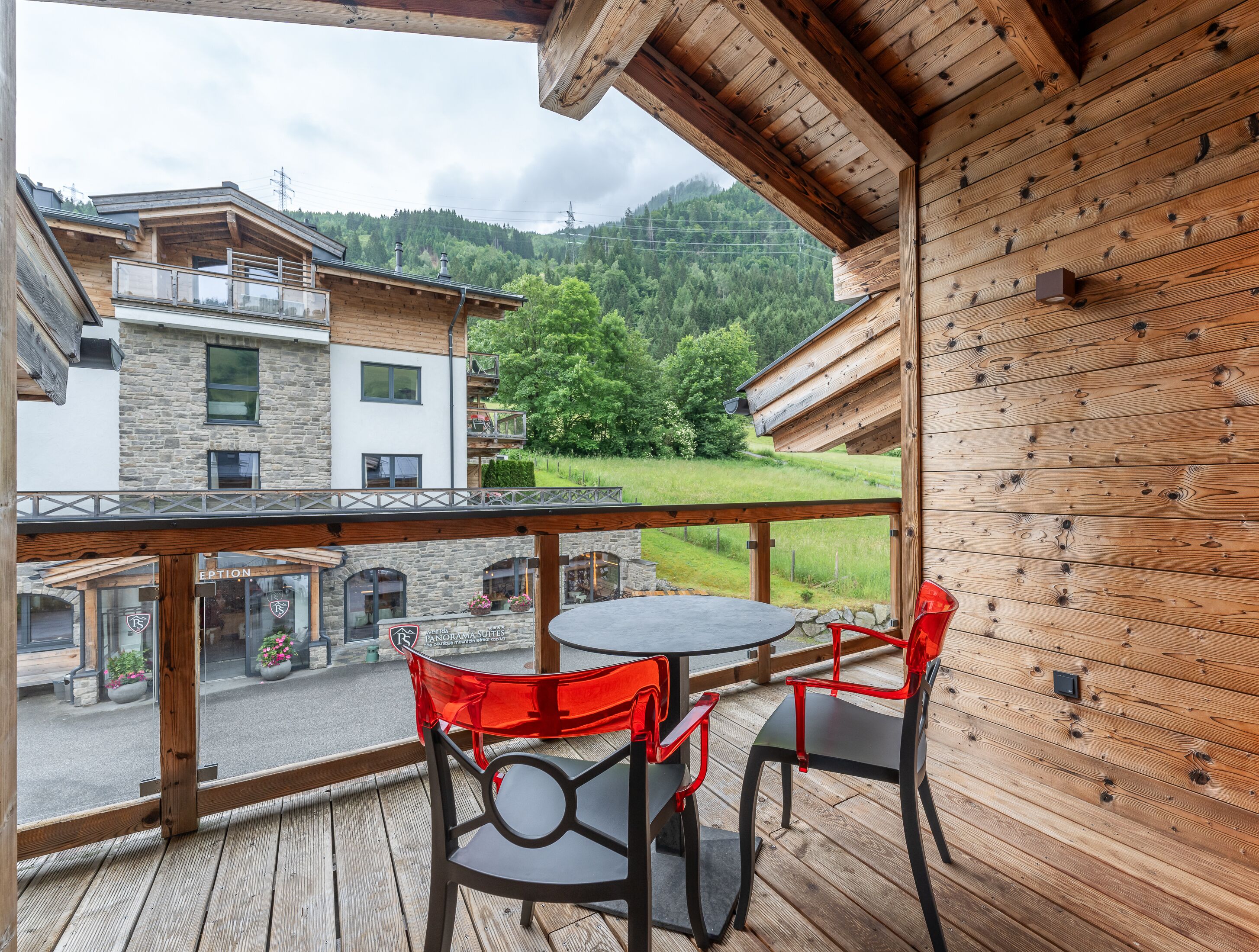 A wooden deck with a round table and red chairs, overlooking a mountain landscape and a building with stone walls.