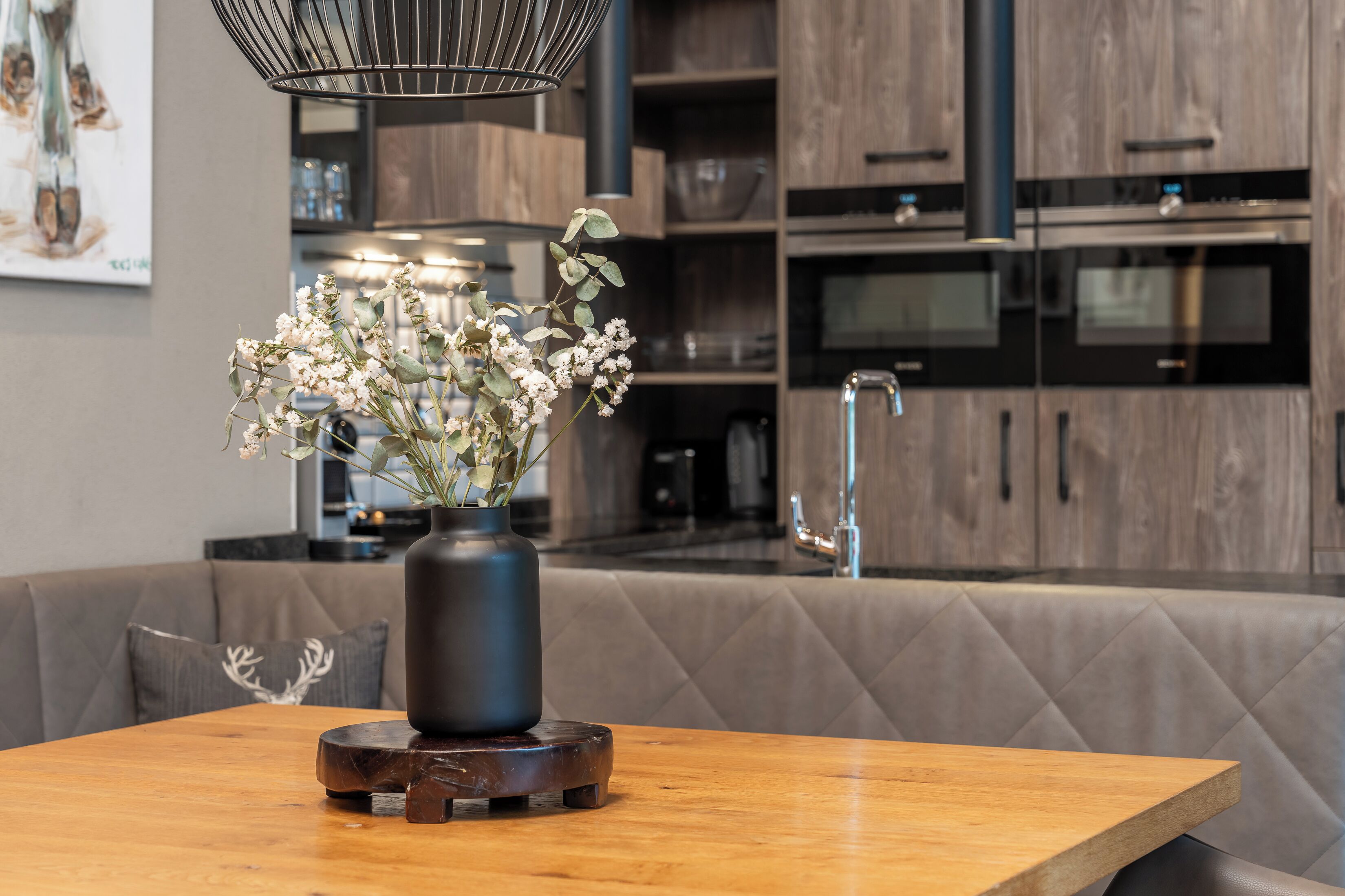 A modern kitchen with wooden cabinets, a built-in oven, and a dining area with a wooden table and a vase of flowers.