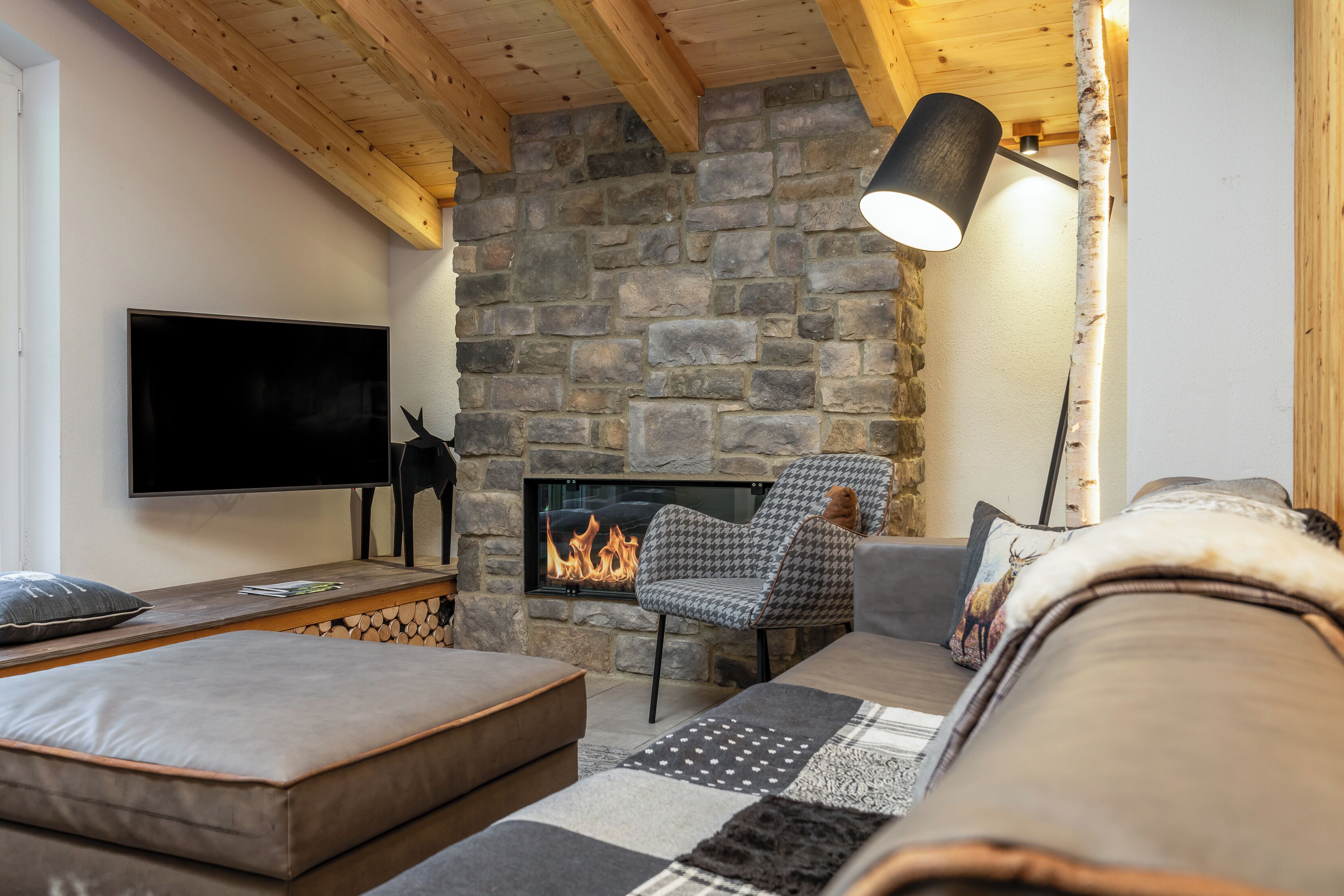 Ein gemütliches Wohnzimmer mit Kamin aus Naturstein, Flachbildfernseher, bequemer Couch und einem gemusterten Teppich.