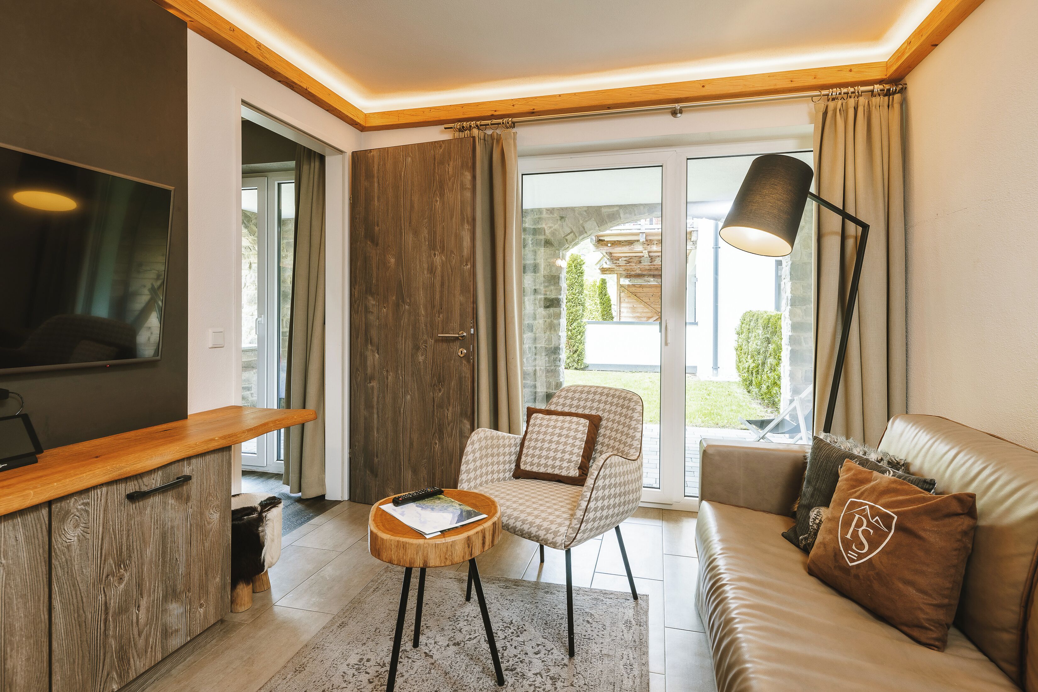 Ein modernes Wohnzimmer mit einer Couch, einem Sessel und einem Couchtisch. Ein Fernseher ist an der Wand befestigt, es gibt eine Lampe und eine Schiebetür zu einem Außenbereich.