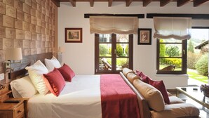 A hotel room with a large bed, a sofa, a table, and a view of the garden through the windows. - Apartamentos Llugarón (Villaviciosa)