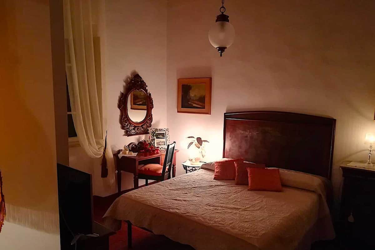 A bedroom with a large bed, a wooden headboard, a desk with a chair, a mirror, and a hanging light fixture.