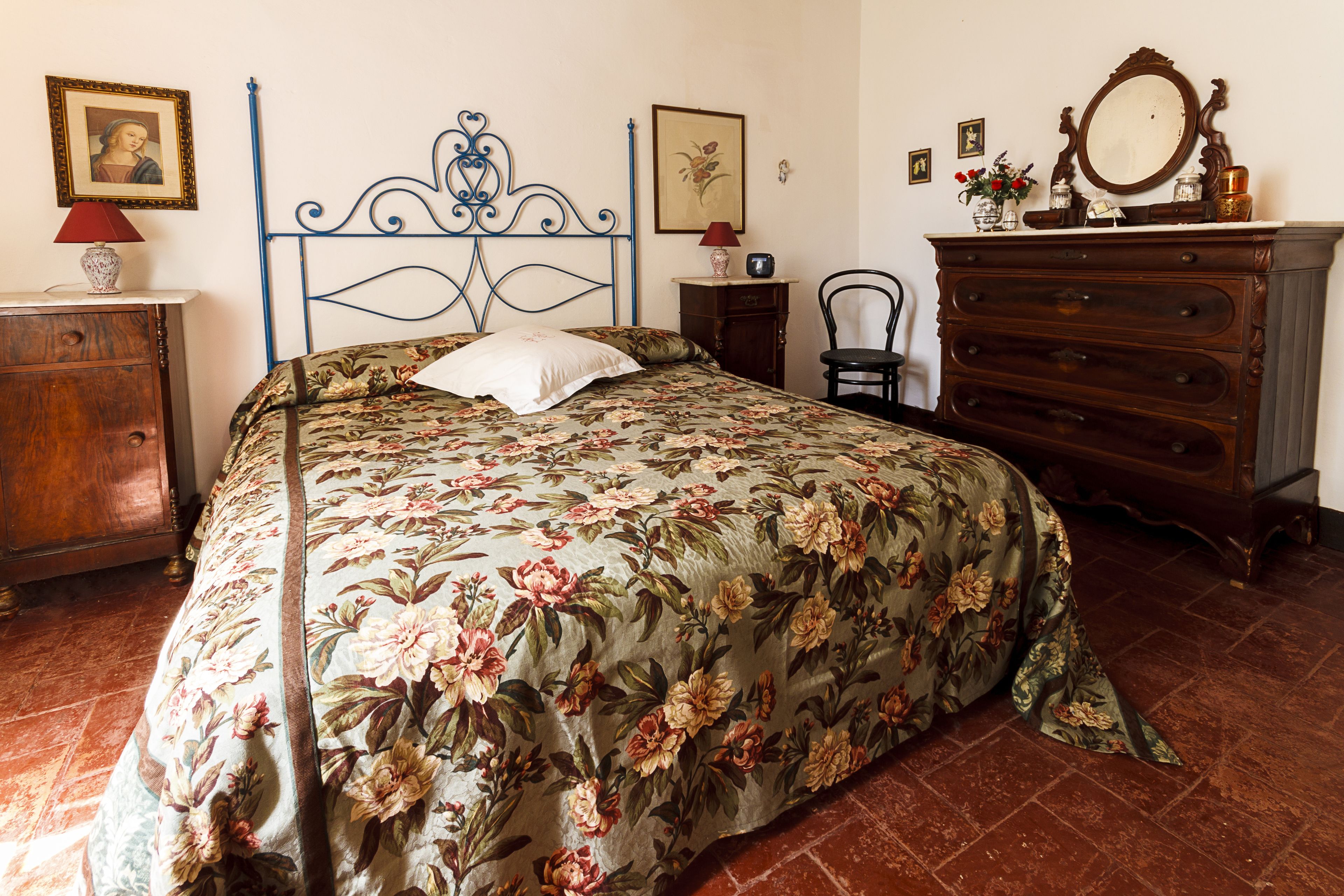 Une chambre à coucher avec une couvre-lit à motifs floraux, une commode en bois, une chaise et une table de chevet avec une lampe.
