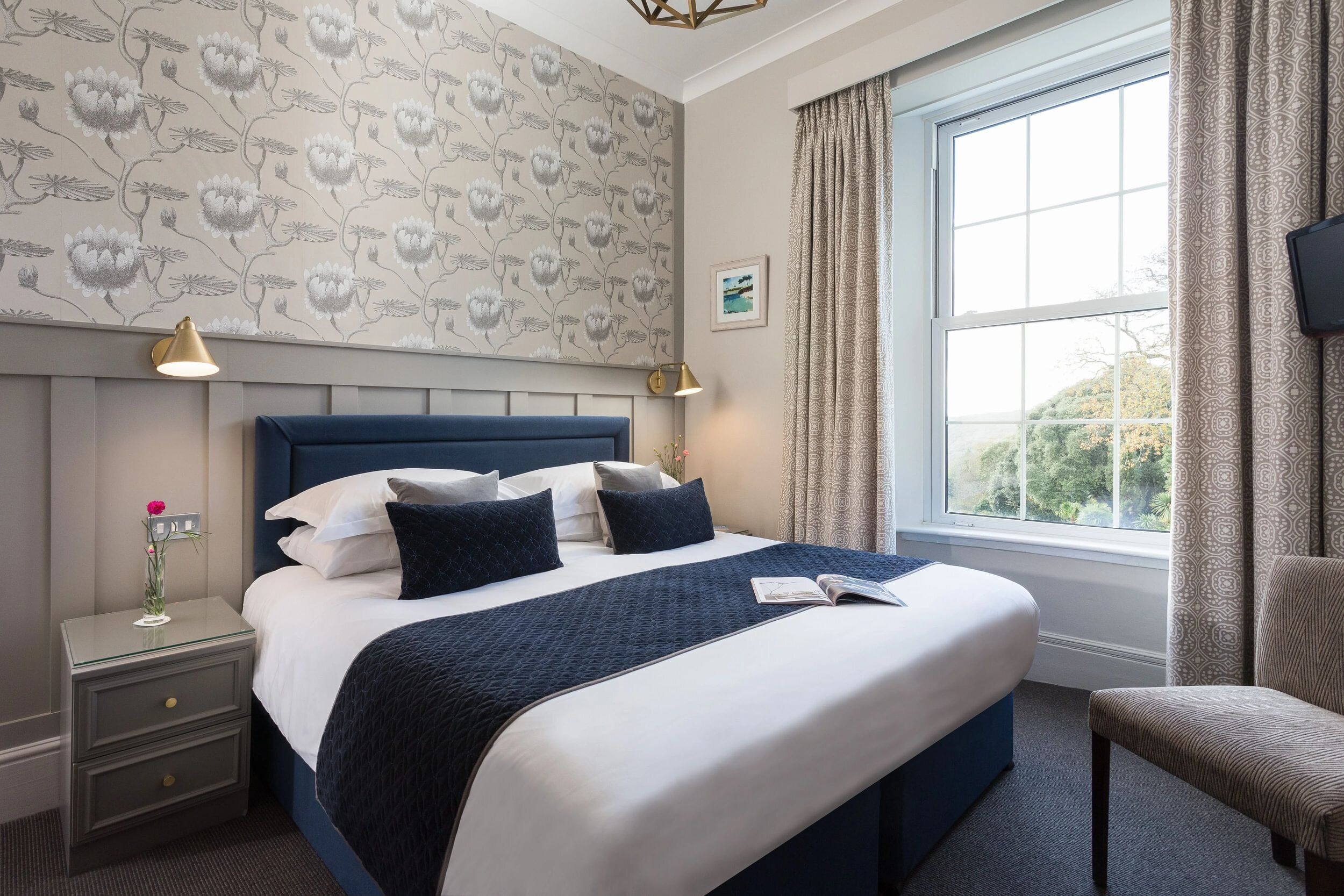 A neatly arranged hotel room with a large bed, bedside lamps, a nightstand with a vase, and a view of the outdoors through a window.
