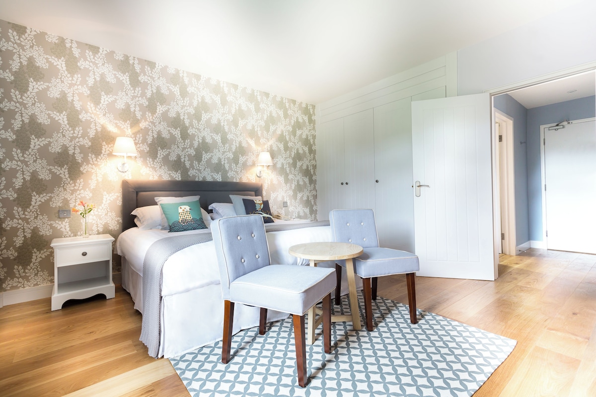 A bedroom with a bed, two chairs, a nightstand, a lamp, and a patterned rug.