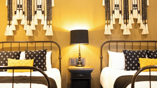 Habitación de hotel con dos camas, cada una con almohadas estampadas, una mesita de noche con lámpara y flecos en la pared.
