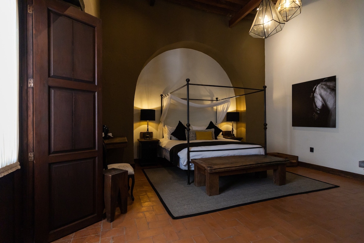 Un dormitorio con una cama con dosel, un banco, una mesita pequeña y una silla. Se observa un amplio arco de entrada y un cuadro colgado en la pared.