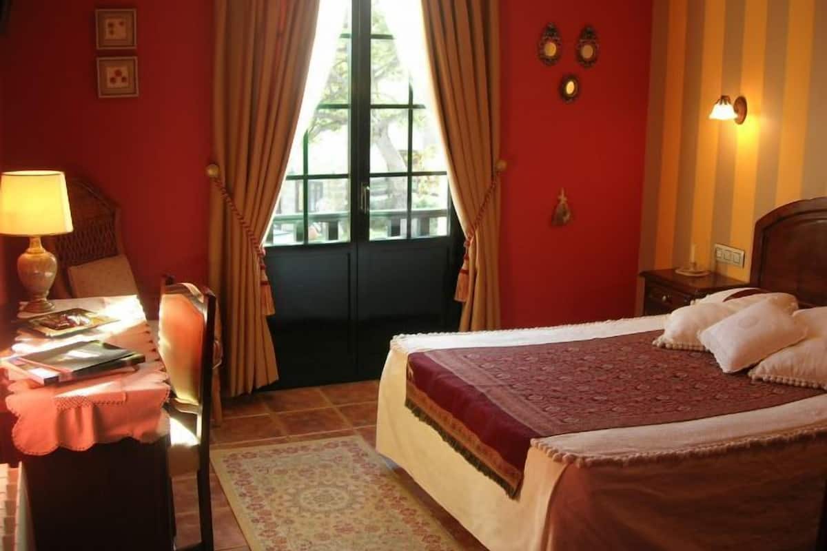 A bedroom with a large bed, a desk with a lamp, a chair, and a window with curtains.