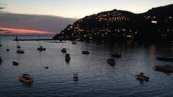 Paisaje costero con barcos en aguas tranquilas, una colina con luces y una zona de embarcadero con gente.