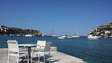 Un puerto deportivo con varios veleros amarrados, un muelle de hormigón con sillas y mesas blancas, y una colina con edificios al fondo.