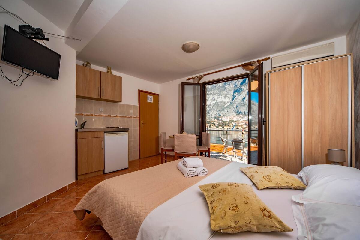 A hotel room with a bed, a desk, a chair, a small kitchenette, and a balcony with a city view.