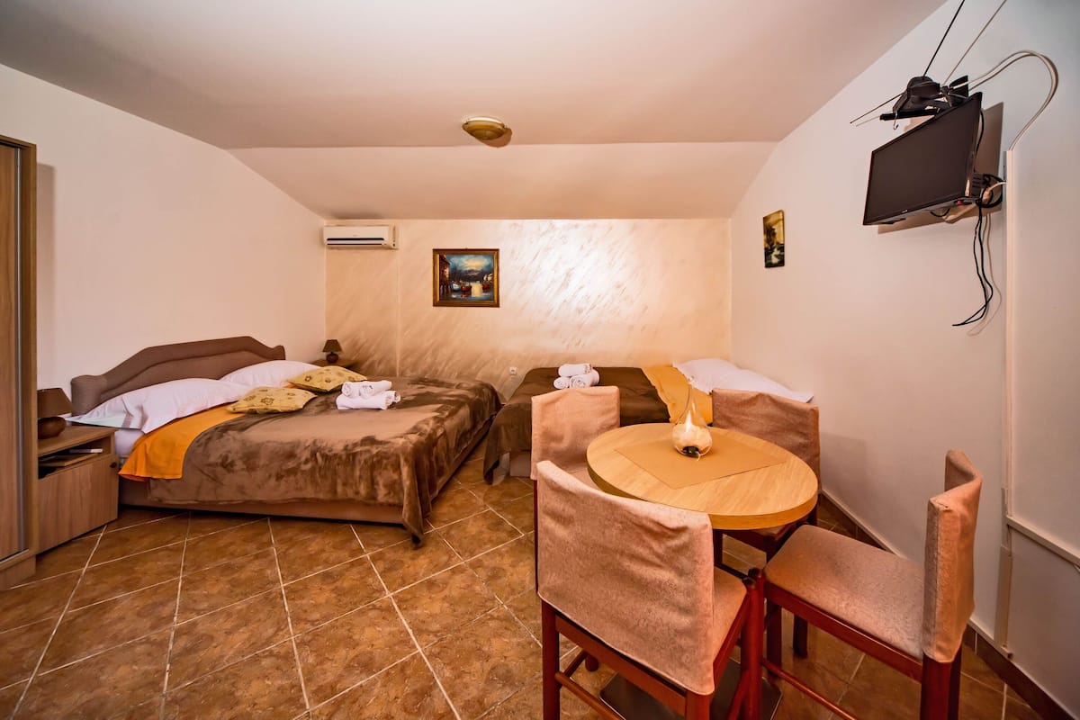 A hotel room with two beds, a dining table with chairs, a TV mounted on the wall, and an air conditioning unit.