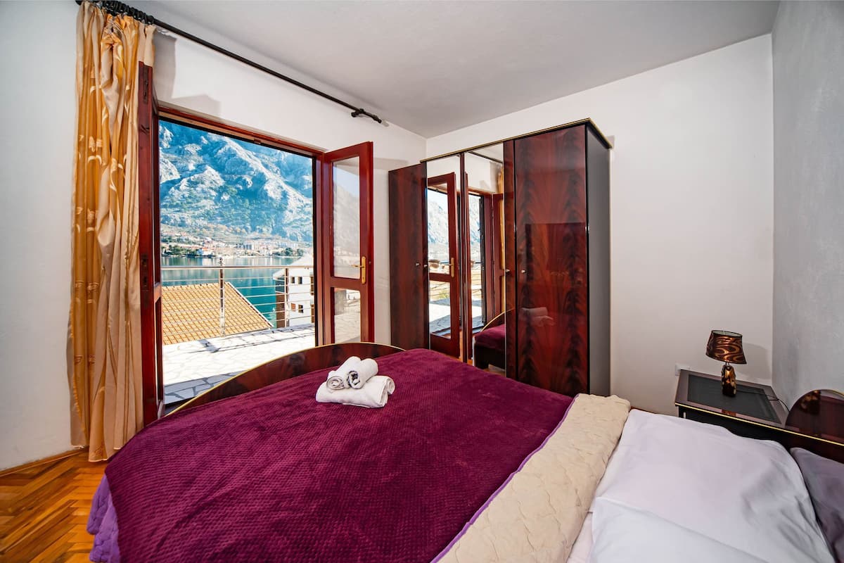 A bedroom with a large bed, a view of mountains and a city through an open door, a wooden wardrobe, and a bedside table with a lamp.