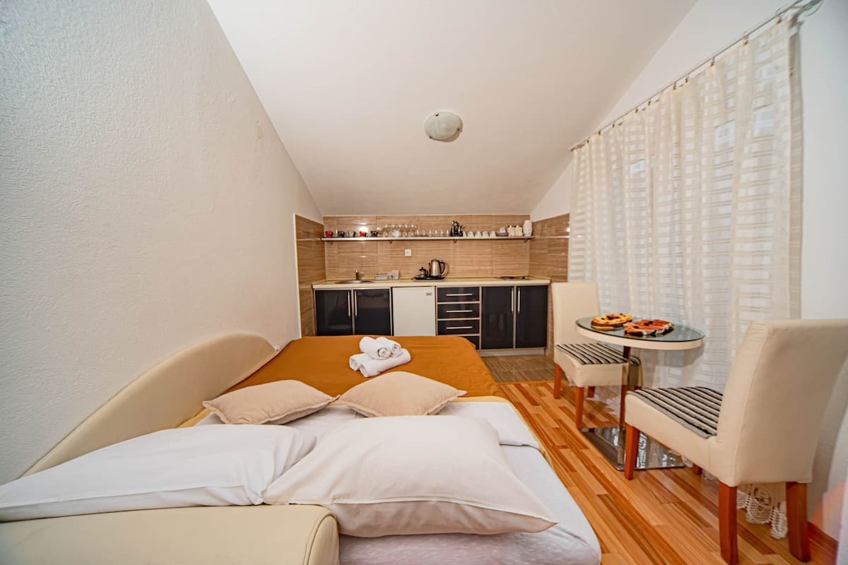 A compact hotel room with a bed, a small table with a chair, a kitchenette, and a bathroom.