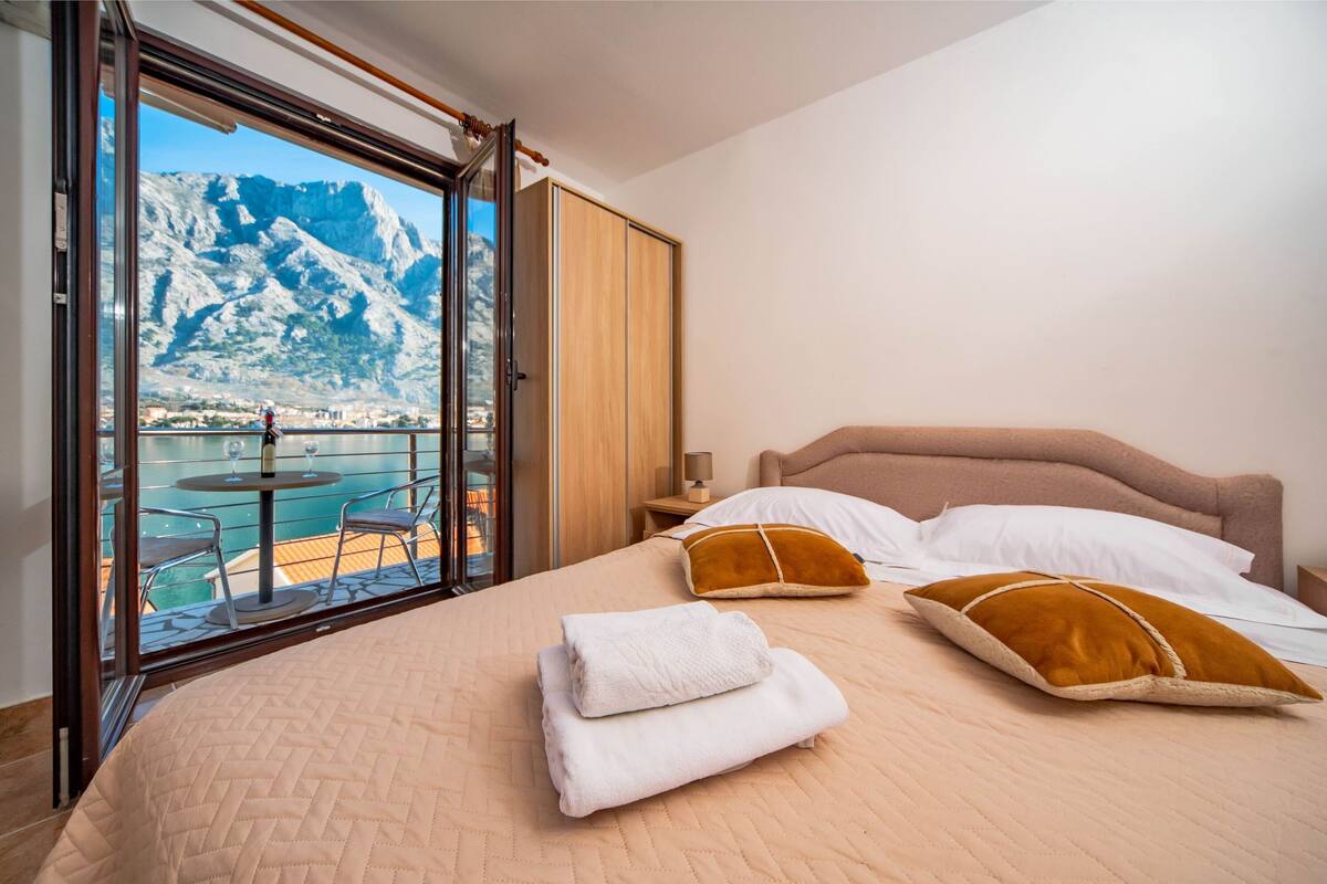 A hotel room with a bed, pillows, a nightstand, and a view of a mountainous landscape through a sliding door.