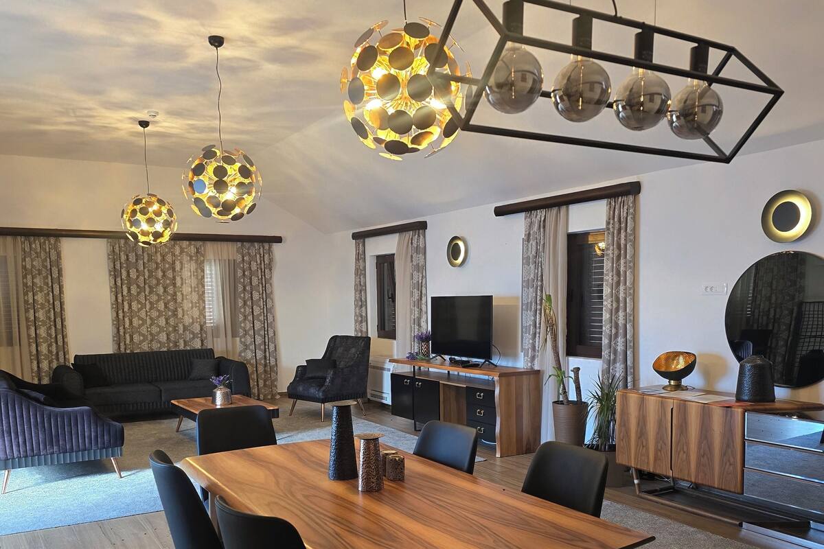 Un salón moderno con una mesa de comedor de madera, sillas negras y un televisor. Se observan dos lámparas colgantes esféricas y una lámpara de techo geométrica.