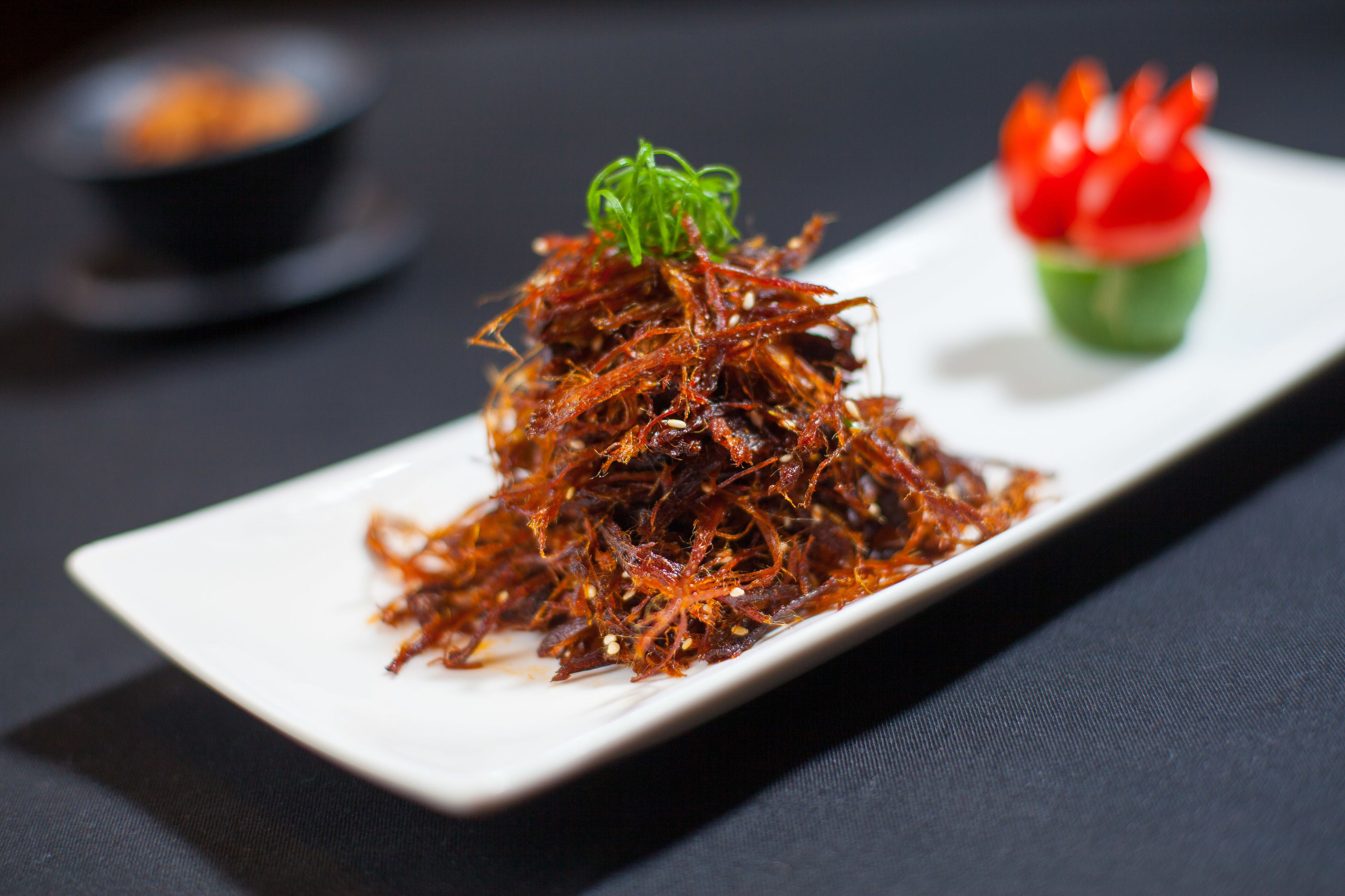 A white rectangular plate with a pile of red, crispy food garnished with green and red elements, placed on a dark surface.