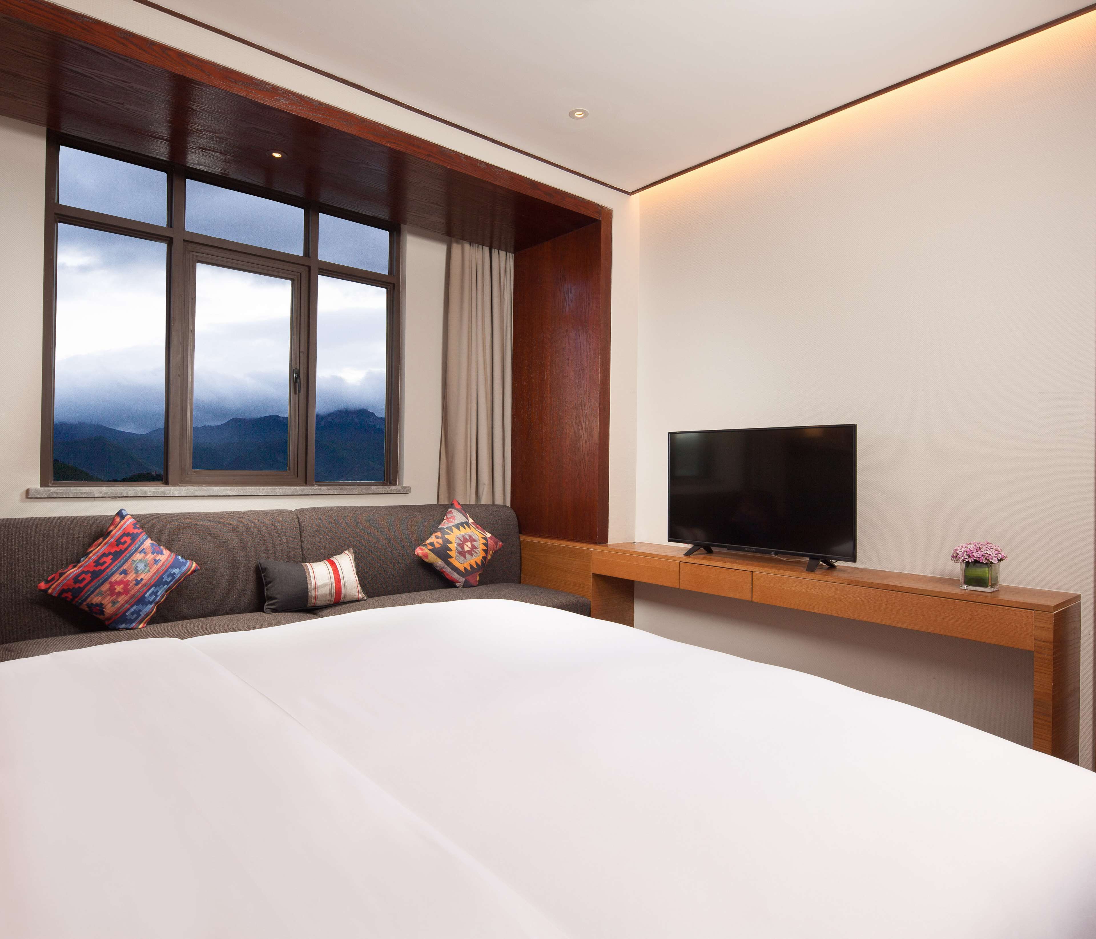 Habitación de hotel moderna con una cama grande, un televisor sobre un mueble de madera y una ventana con vistas a la montaña.