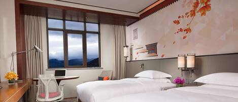 Une chambre d’hôtel avec deux lits, un bureau avec un ordinateur portable, une chaise, une lampe de bureau et une vue sur les montagnes par la fenêtre.