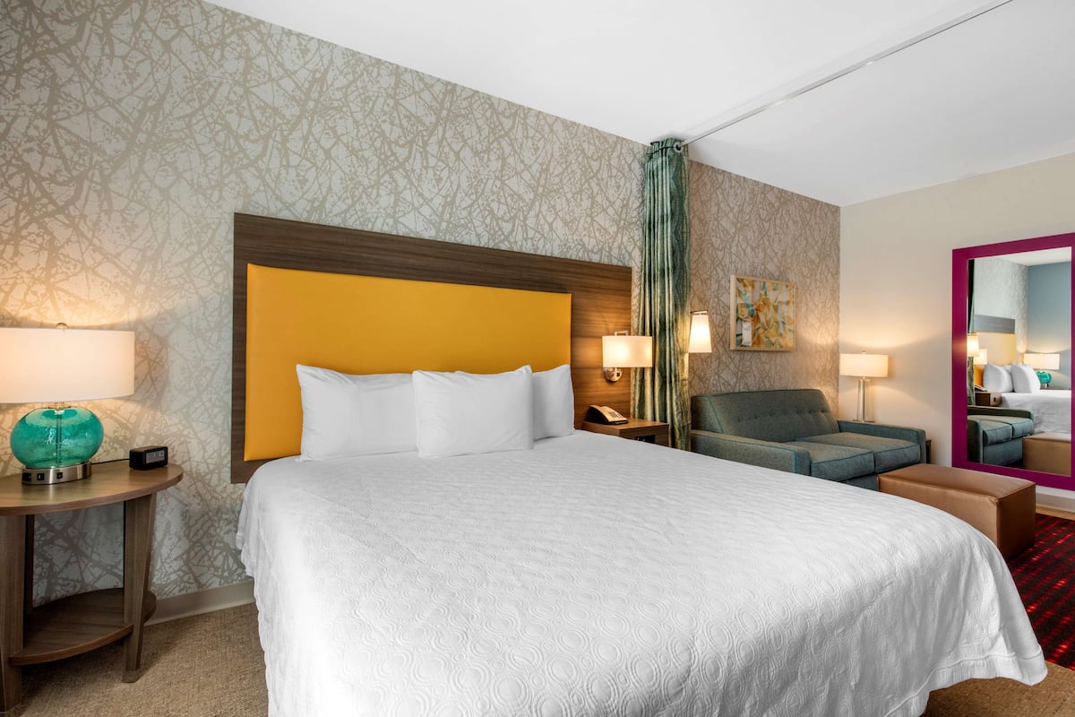 A hotel room with a large bed, a nightstand, a chair, a mirror, and a patterned wallpaper.