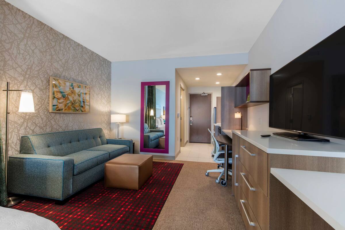 A modern hotel room with a sofa, a small table, a TV, and a kitchenette.