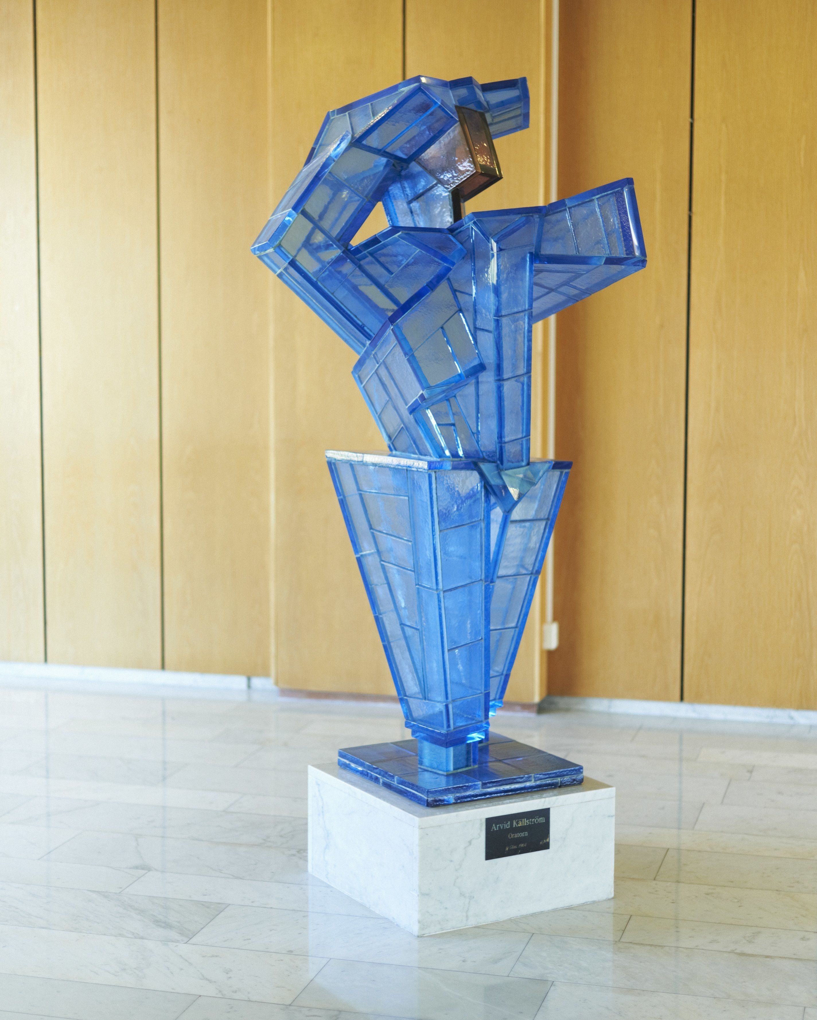 A blue glass sculpture on a white pedestal in a lobby with wooden paneling and a tiled floor.