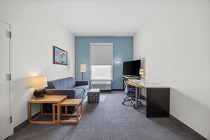 A compact hotel room with a sofa, a small table, a desk with a chair, a TV, and a window with blinds. - Home2 Suites By Hilton Maumee Toledo (Maumee)