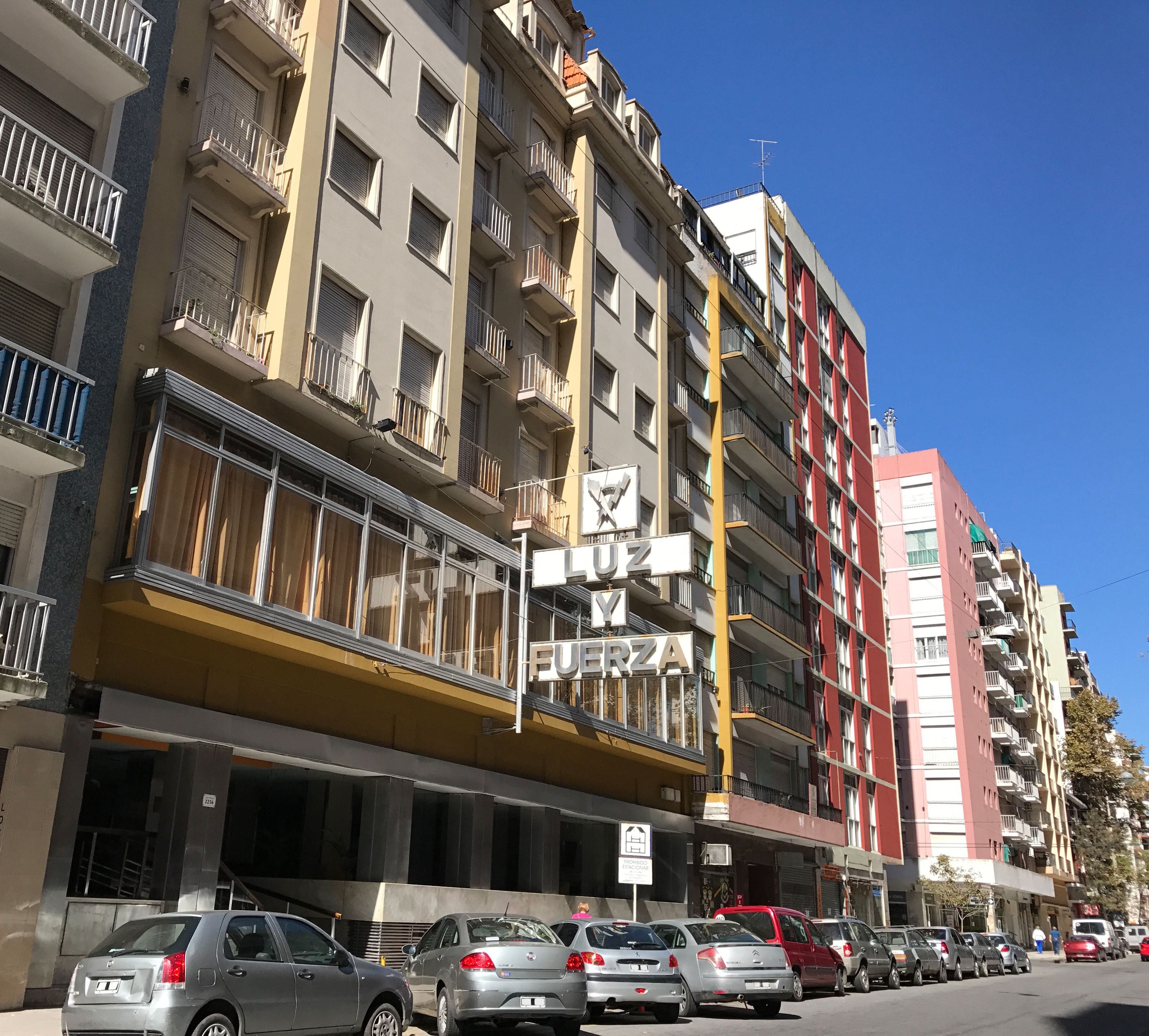 Un edificio de varios pisos con balcones, un cartel que dice 'Y & FUERZA' a la vista, y varios coches aparcados en la calle.