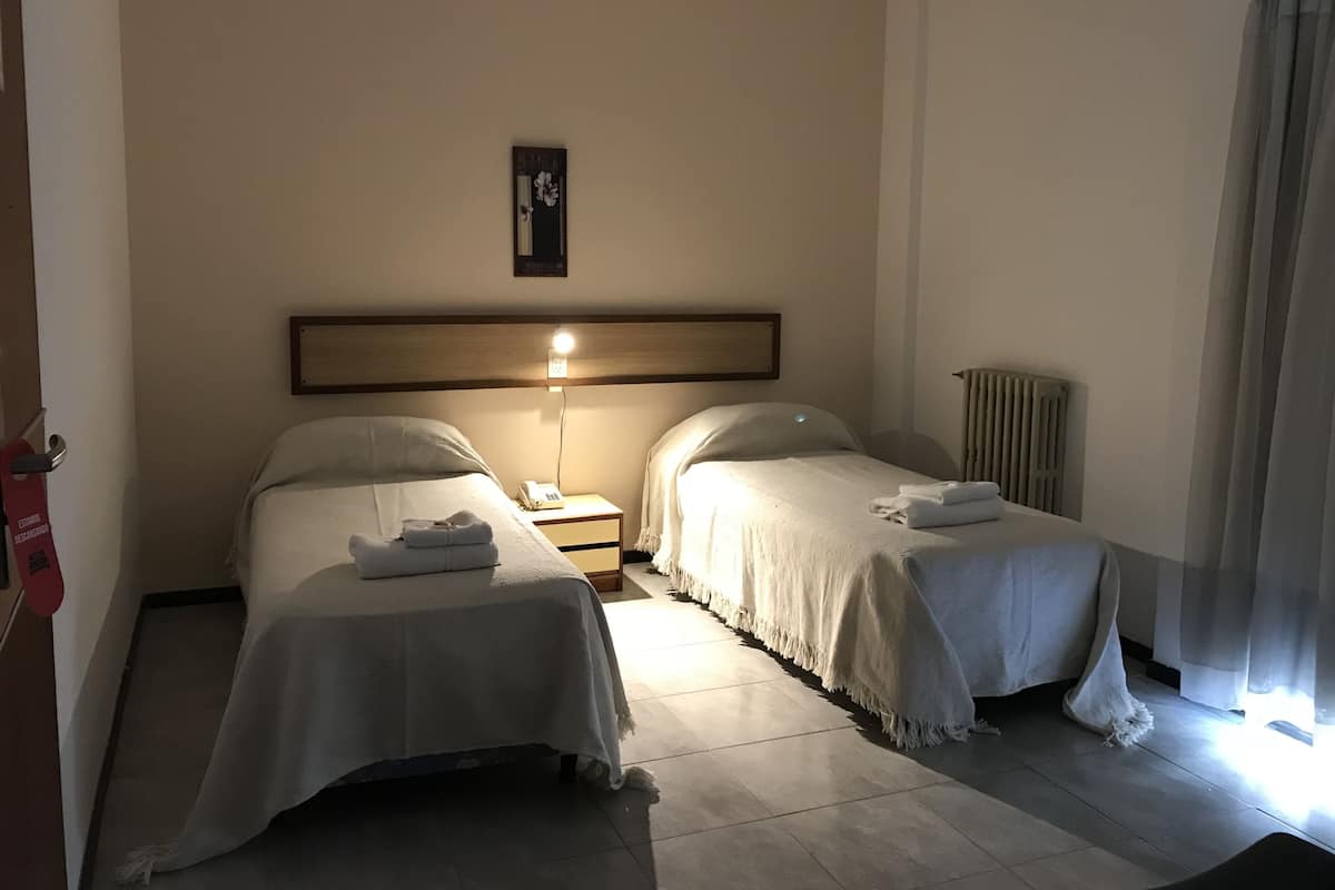 Habitación de hotel con dos camas individuales, una mesita de noche con lámpara, un termostato y un cuadro en la pared.