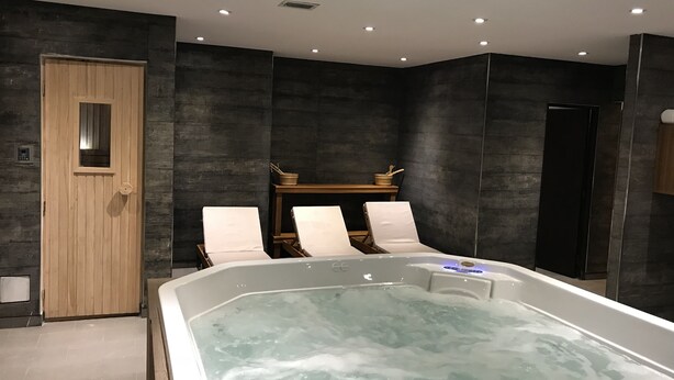 Un spa moderno con una bañera de hidromasaje grande, dos sillones de descanso, una mesa de madera con un cuenco y palillos, y una pared oscura con una puerta de madera.