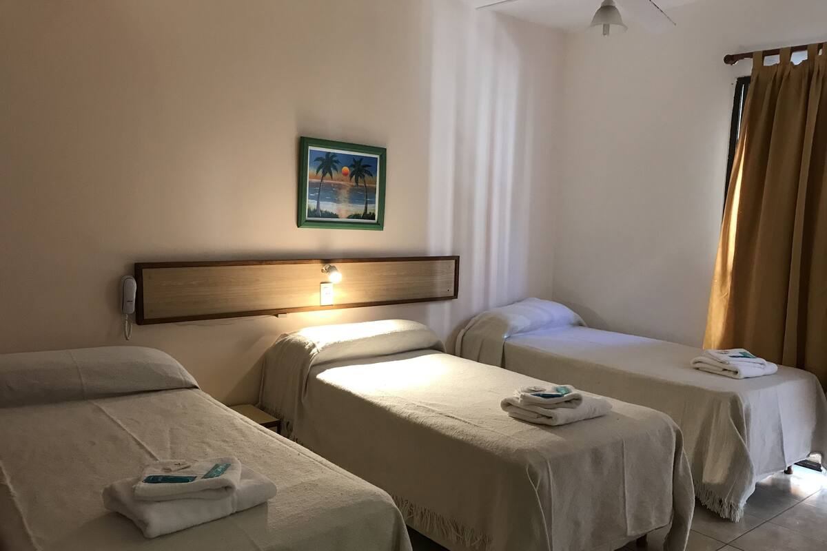 Habitación de hotel con dos camas individuales, una mesita de noche con lámpara, un cuadro en la pared y una ventana con cortinas.