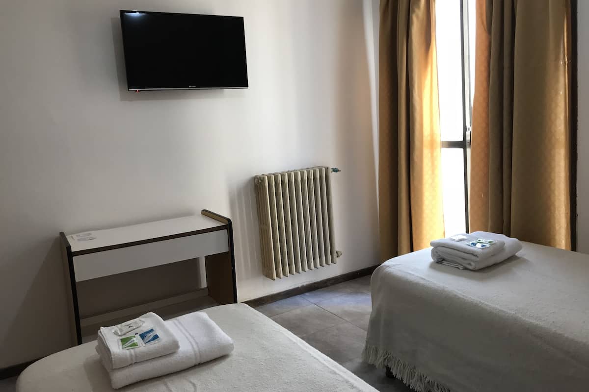 Habitación de hotel con cama, mesita de noche, televisor montado en la pared, radiador y ventana con cortinas.