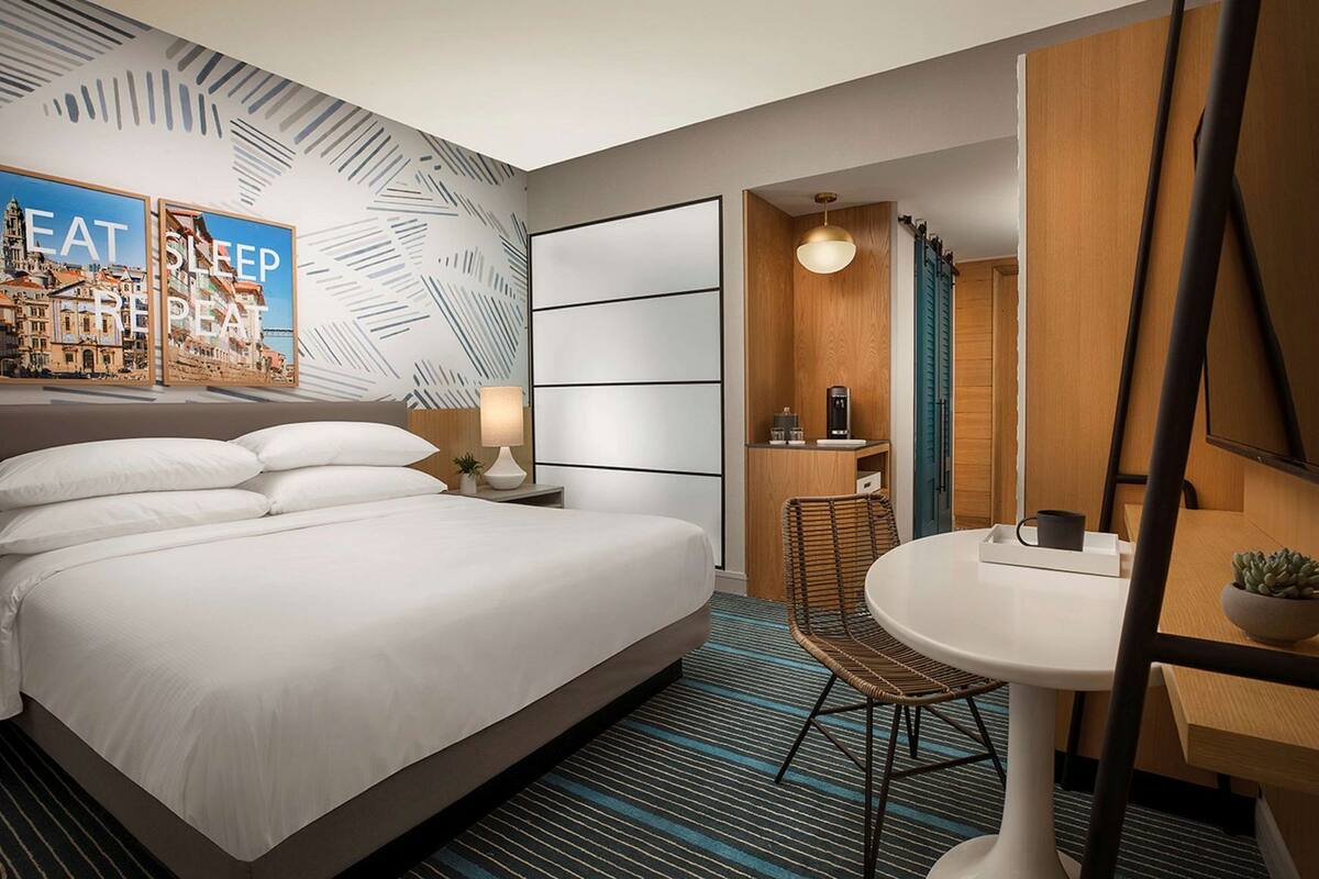 A hotel room with a bed, a desk, a chair, a round table, a mirror, and a wall with a leaf pattern and a poster.