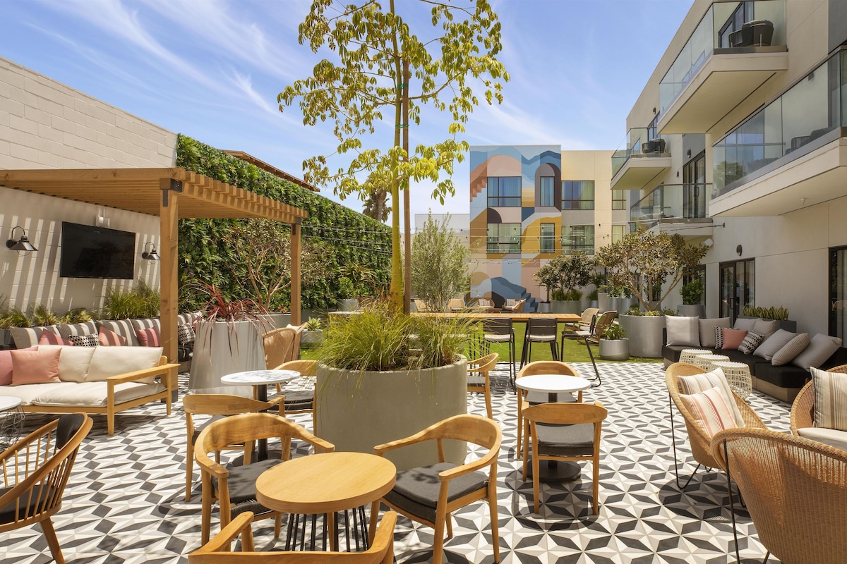 An outdoor patio area with seating arrangements, a pergola, and a view of a modern building.