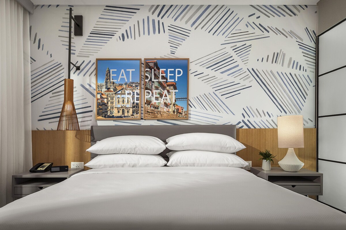 A modern hotel room with a large bed, two bedside tables, a wall-mounted lamp, and a poster with the text 'EAT SLEEP REPEAT'.