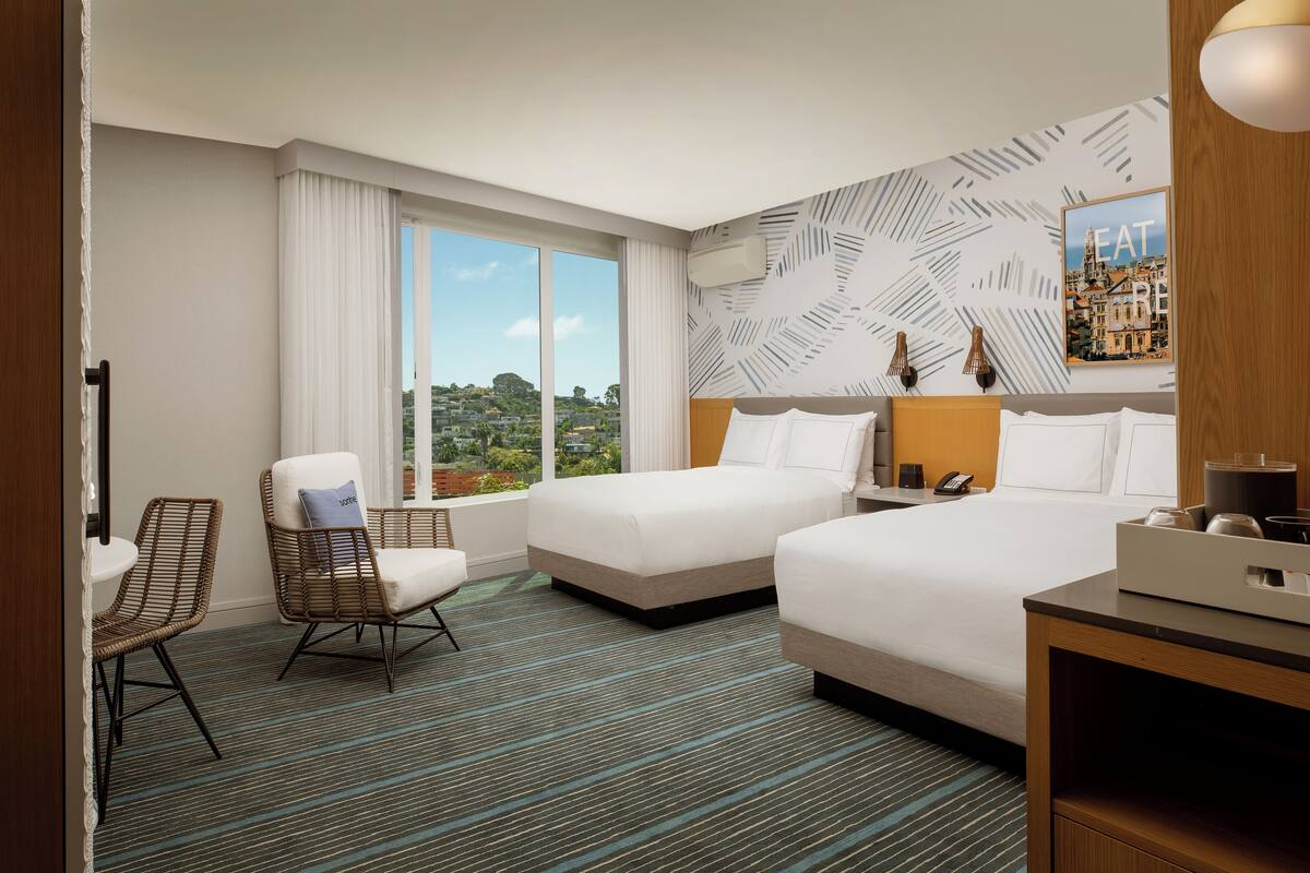A hotel room with two beds, a large window with a view, a chair, and a nightstand.