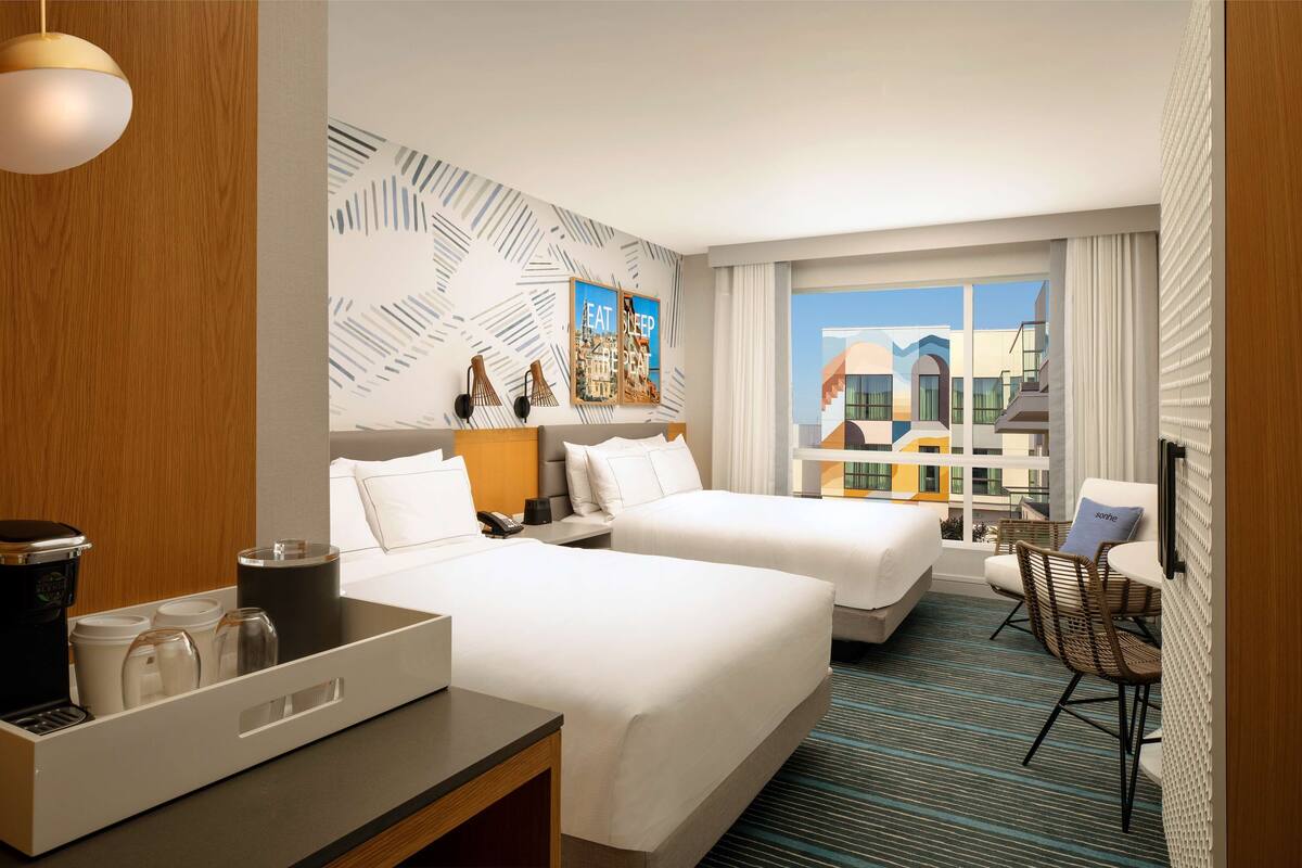 A hotel room with two beds, a desk with a coffee maker, a chair, and a large window offering a city view.
