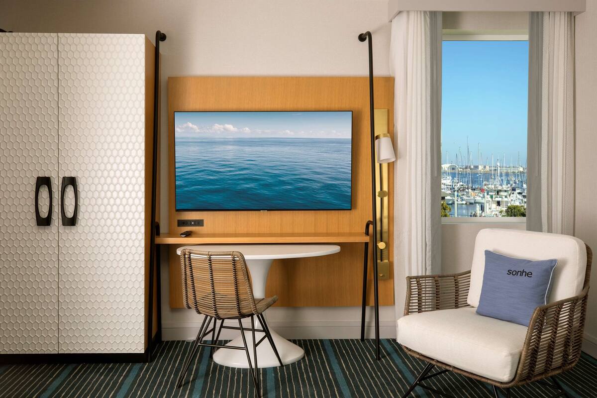 A hotel room with a TV, a dining table, a chair, a sofa, a wardrobe, and a view of a marina.
