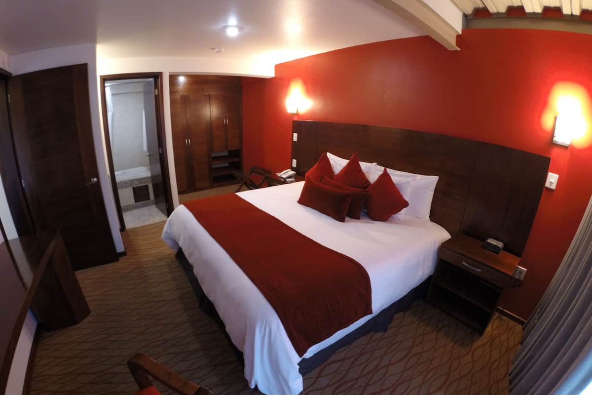 A hotel room with a large bed, a wooden wardrobe, a desk with a chair, and a bathroom visible through an open door.