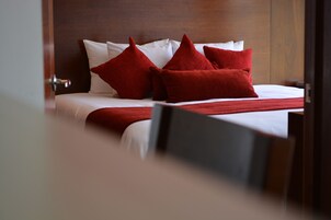 Cama bem arrumada com lençóis brancos e almofadas vermelhas, cabeceira de madeira e puxador de porta visível.