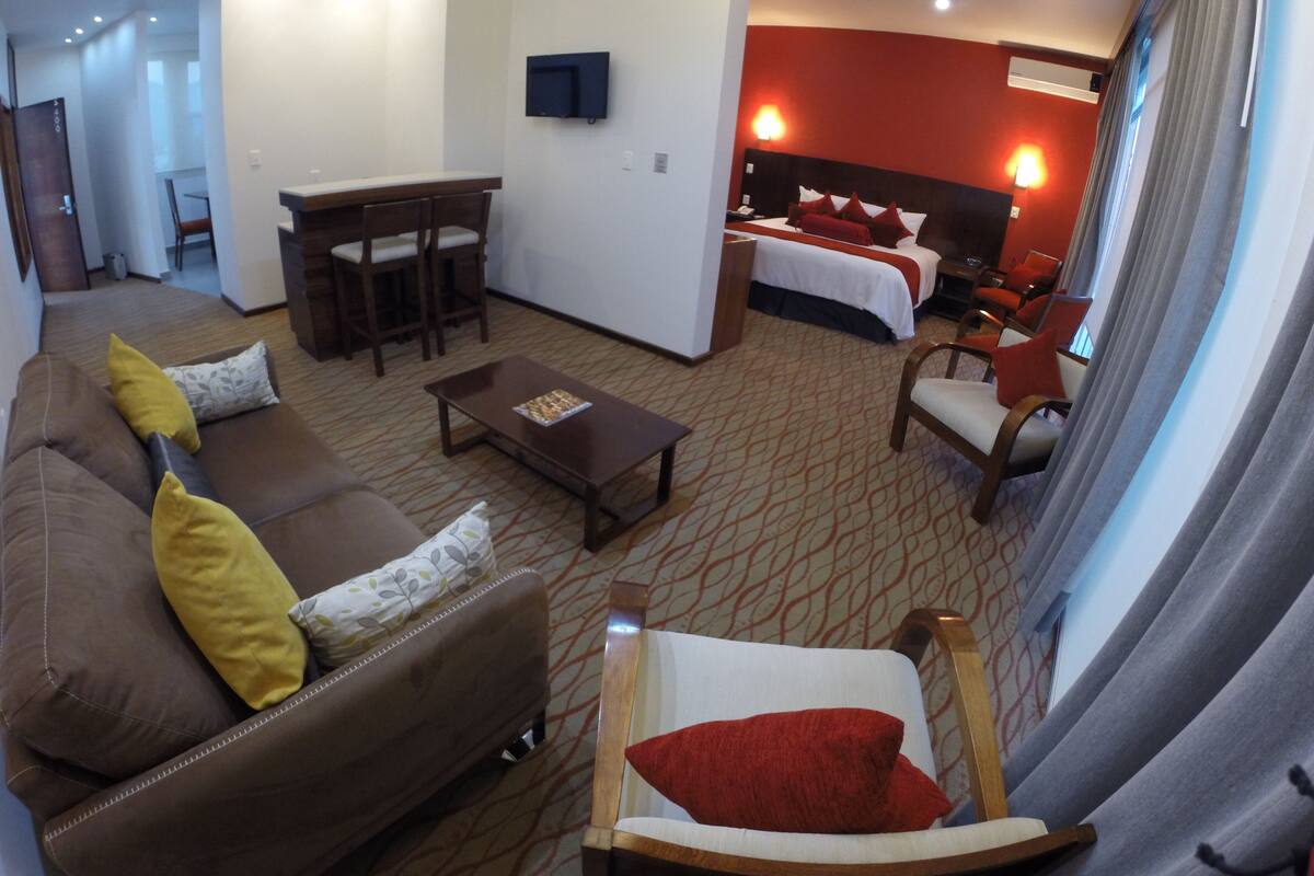 A hotel room with a living area, a dining table, a sofa with cushions, a chair, and a bed with red pillows.